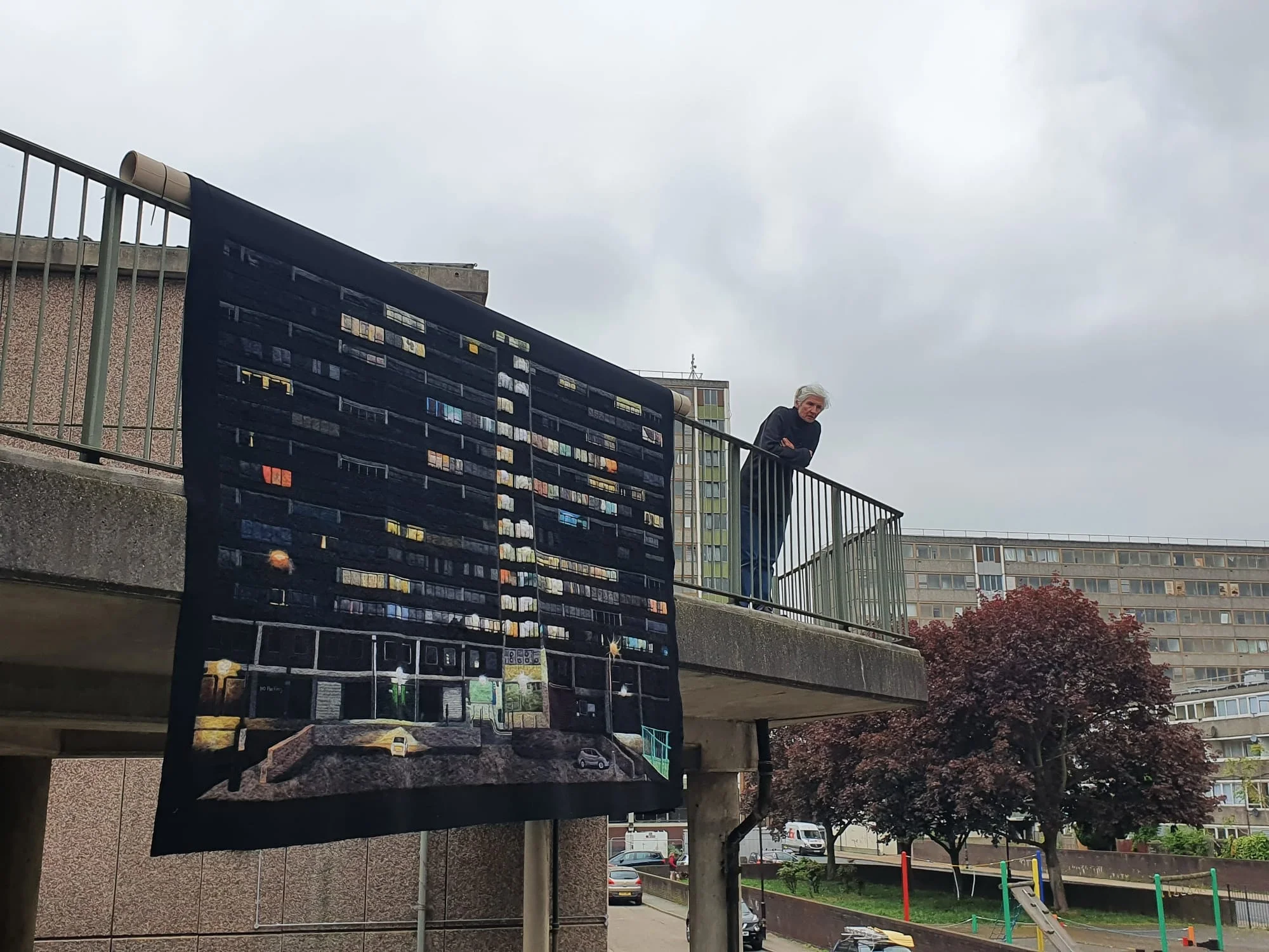 a textile piece depicting public housing hangs over a balcony while the artist leans on the railing nearby